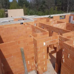 Wznoszenie ścian parteru z czerwonej ceramiki poryzowanej, widoczne spoiny wypełnione pianką montażową, w tle las i fragment budynku.