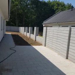 Nowoczesny, szary, betonowy płot modułowy z przęsłami ułożonymi poziomo, oddzielający posesję od sąsiada, widziany z perspektywy brukowanej ścieżki.