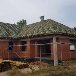 Surowy stan domu z czerwonej cegły z widocznym drewnianym szkieletem dachu pokrytym zieloną membraną, komin z cegły oraz rusztowania.