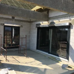 Surowy stan budynku w trakcie budowy z zamontowanymi oknami przesuwnymi z roletami zewnętrznymi i oknem stałym, widoczne rusztowanie i materiały budowlane.