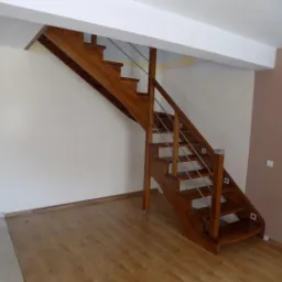 Klasyczne schody drewniane z metalowymi relingami, polakierowane na wysoki połysk, w jasnym wnętrzu z panelami podłogowymi.