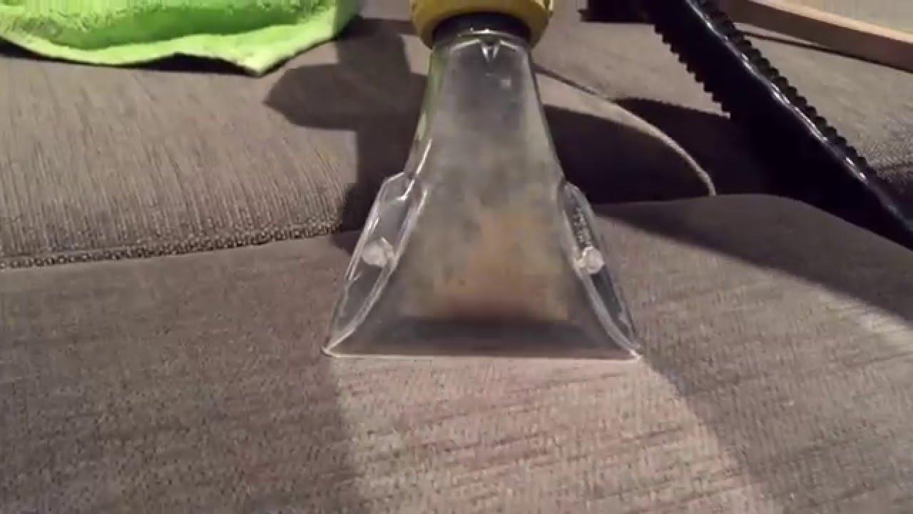 Czyszczenie tapicerki kanapy za pomocą odkurzacza piorącego z przezroczystą dyszą, widoczne zabrudzenia w dyszy oraz fragment wyczyszczonej powierzchni.