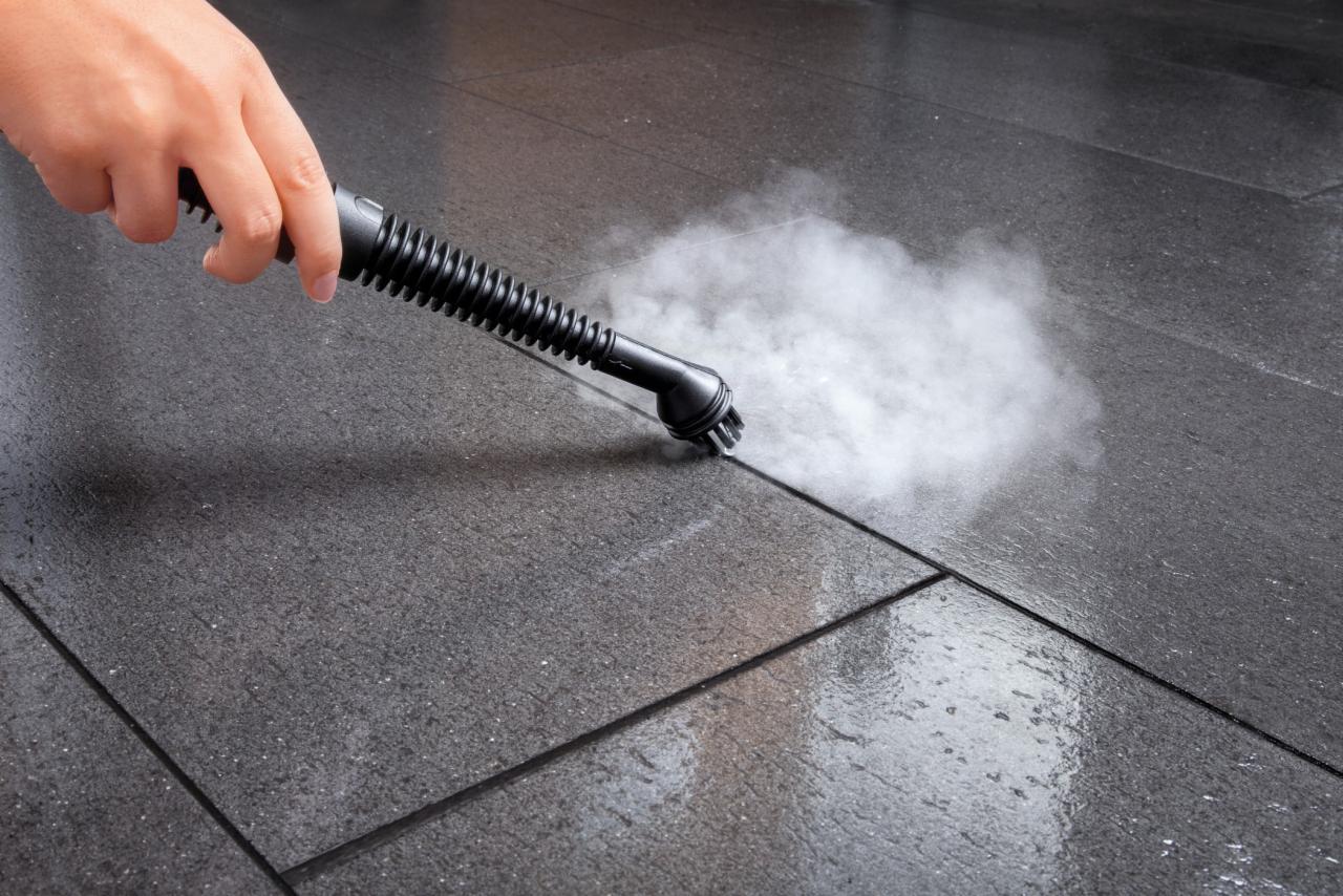 Czyszczenie parowe ciemnych płytek podłogowych w Toruniu za pomocą parownicy z dyszą szczotkową, widoczna para wodna.