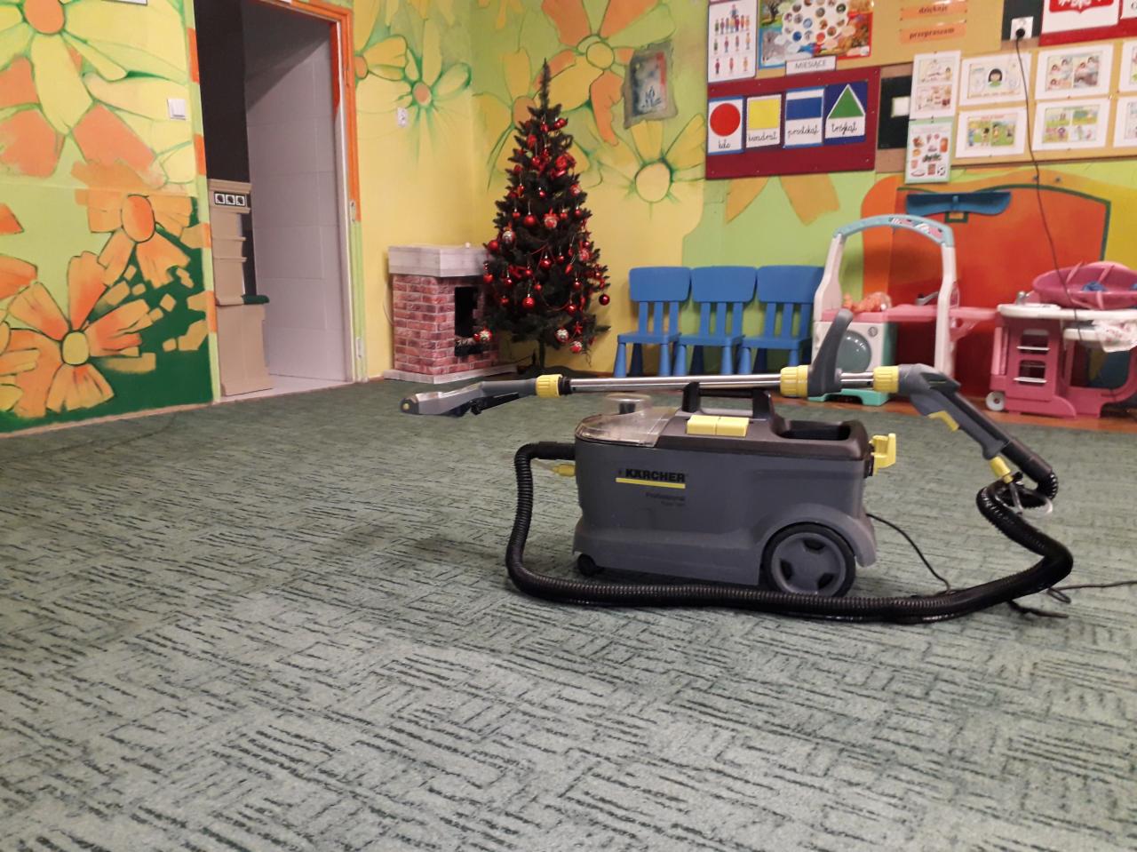Profesjonalny odkurzacz piorący Karcher na tle zielonej wykładziny w pokoju dziecięcym z malowidłami na ścianach, atrapą kominka i choinką.