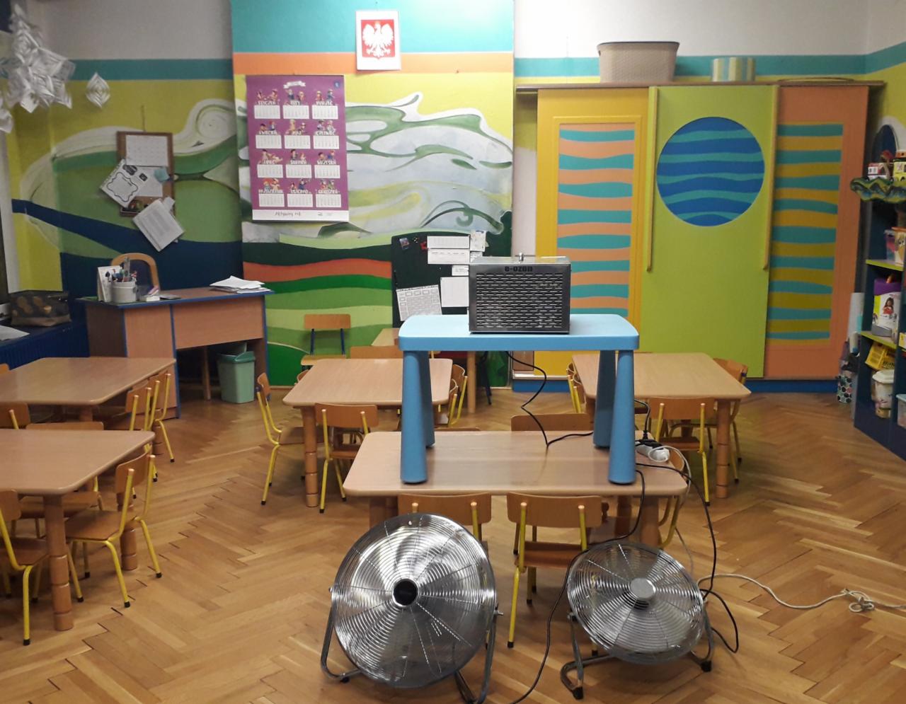 Sala przedszkolna z pomalowanymi ścianami, drewnianą podłogą, stolikami i krzesłami, z ustawionym na środku niebieskim stolikiem, na którym stoi czarny ozonator, a na podłodze dwa metalowe...