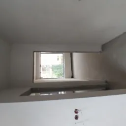 Wnętrze w trakcie prac wykończeniowych: ściany i sufit pokryte świeżym, jasnoszarym tynkiem, widoczne puszki elektryczne bez osprzętu, okno z widokiem na zieleń.