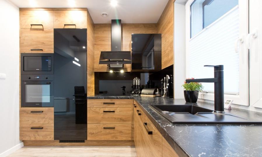 Nowoczesna kuchnia z drewnianymi frontami szafek, czarnymi elementami wykończenia, marmurowym blatem i oknem z żaluzjami plisowanymi.