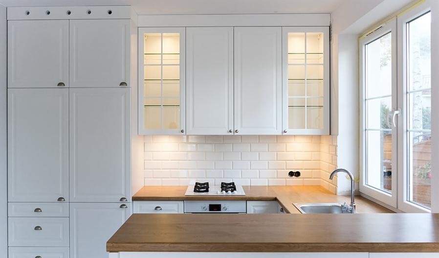 Jasna kuchnia z białymi szafkami, drewnianym blatem i białymi płytkami metra na ścianie, oświetlona ciepłym światłem spod szafek.