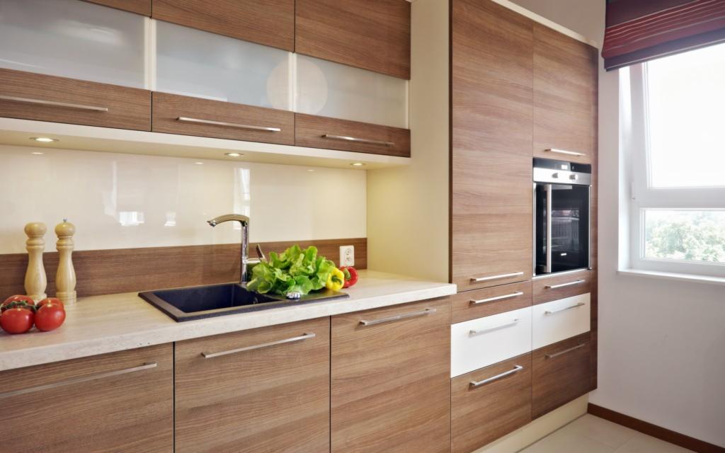 Nowoczesna kuchnia z drewnianymi frontami, przeszklonymi szafkami górnymi i blatem z ciemnym zlewozmywakiem, uzupełniona świeżymi warzywami i przyprawami, oświetlona światłem dziennym z okna.