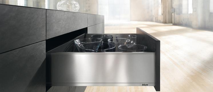 Otwarta szuflada z metalowym frontem, wypełniona szklanymi miseczkami, w nowoczesnej kuchni z ciemnymi, matowymi frontami szafek. W tle jasna podłoga i okno w industrialnym wnętrzu.