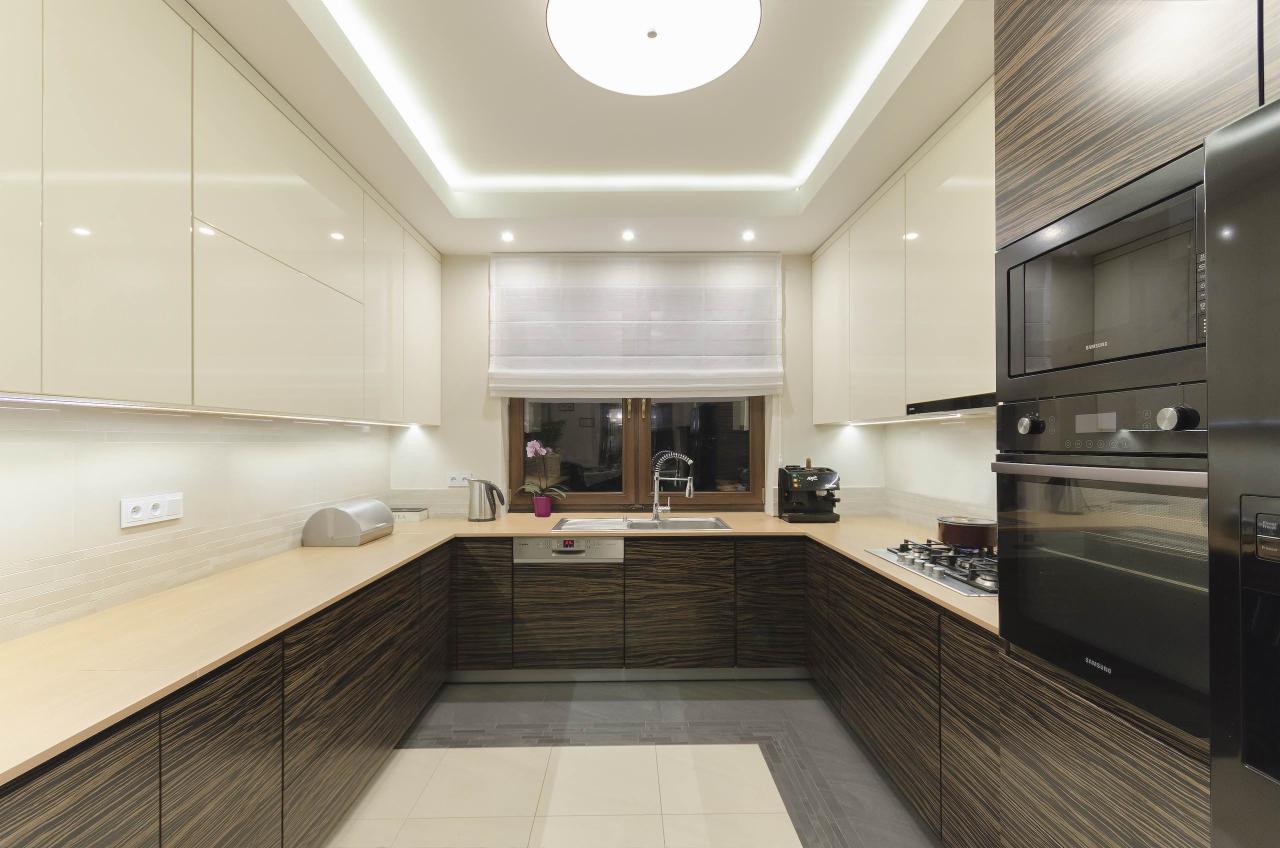 Nowoczesna kuchnia w kształcie litery U z białymi górnymi szafkami i dolnymi szafkami z ciemnego drewna ze wzorem zebry, oświetleniem podszafkowym i centralnym, widok na okno z roletą rzymską.