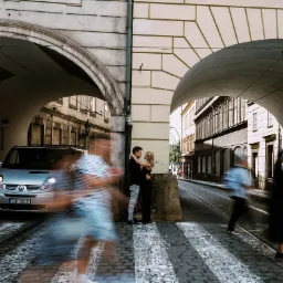 Para obejmująca się na tle zabytkowej ulicy z przejściem dla pieszych i dwoma łukami, z rozmytymi postaciami przechodniów i zaparkowanym samochodem w tle.