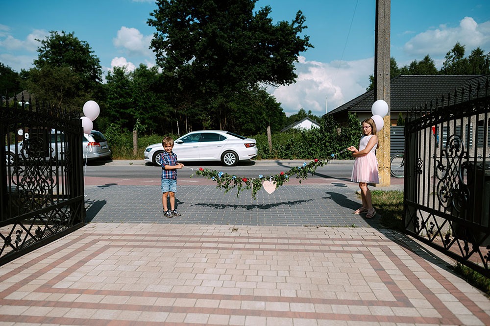 Chłopiec i dziewczynka trzymają girlandę z kwiatów i serca przed bramą, w tle samochody i budynki.