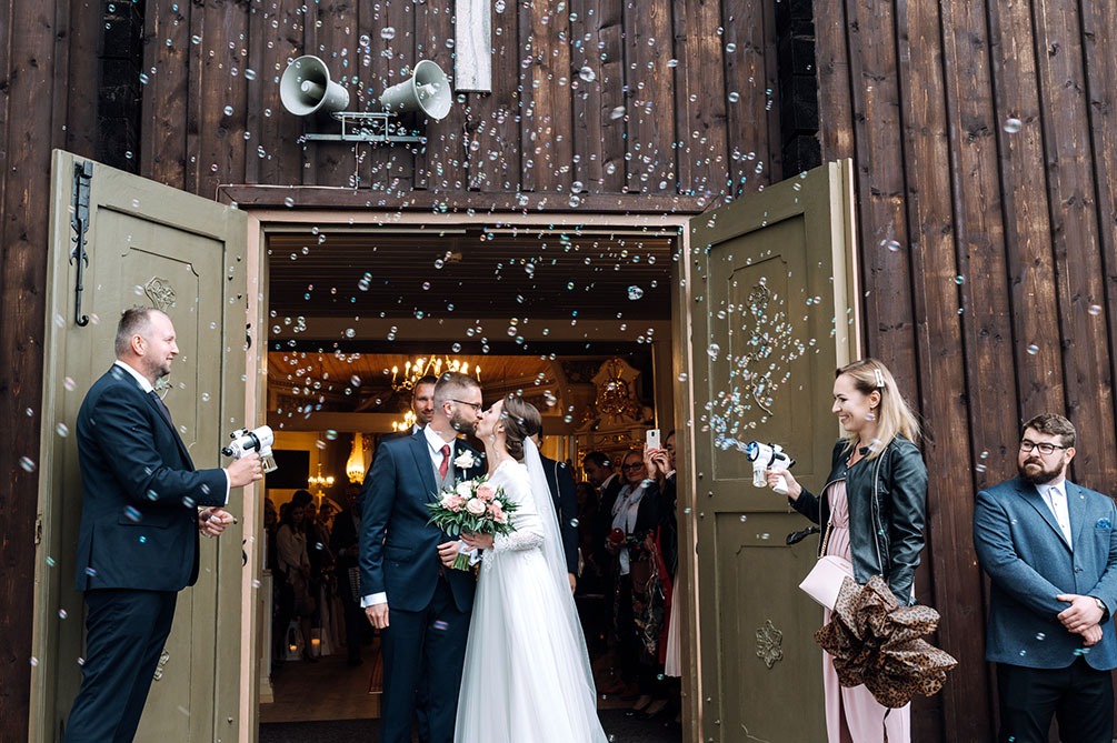 Nowożeńcy całują się na tle drewnianego kościoła, otoczeni bańkami mydlanymi wystrzeliwanymi z pistoletów przez gości weselnych.