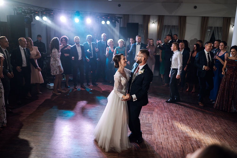 Para młoda tańczy pierwszy taniec na parkiecie w otoczeniu gości weselnych, oświetlona reflektorami. Panna młoda w sukni z koronkową górą i tiulowym dołem, pan młody w granatowym garniturze...