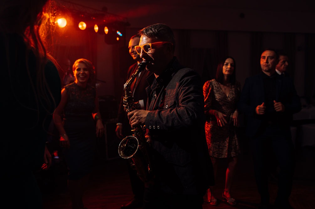 Saksofonista w pomarańczowych okularach gra na saksofonie podczas wydarzenia, w tle tańczący goście w czerwonym oświetleniu.