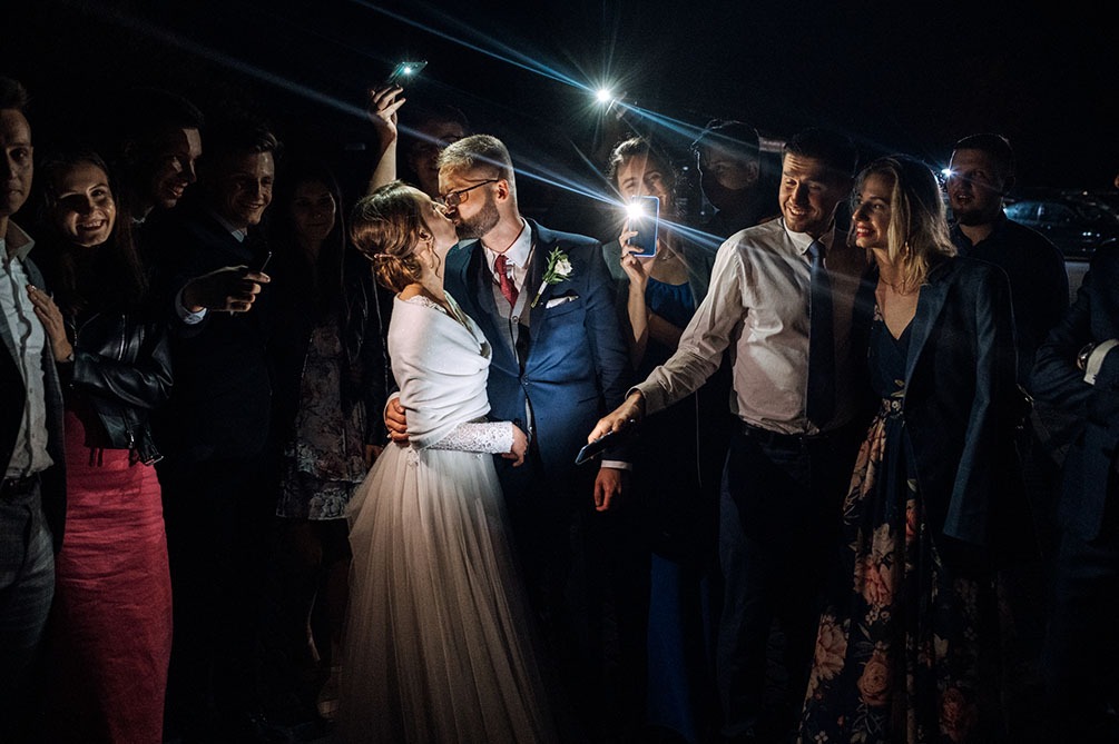 Pocałunek nowożeńców w otoczeniu gości weselnych, oświetlonych smartfonami, panna młoda w białej sukni z koronkowymi rękawami i narzutką, pan młody w granatowym garniturze z bordowym krawatem.