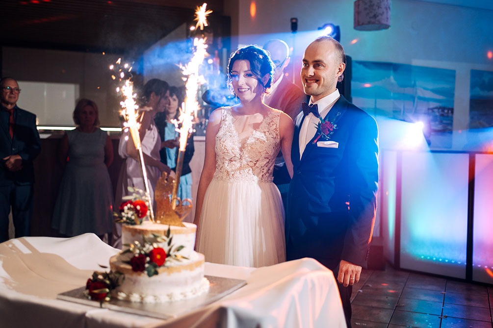 Uśmiechnięta para młoda w otoczeniu gości i tortu weselnego z zimnymi ogniami, on w ciemnym garniturze z butonierką, ona w białej sukni z koronkową górą, tło rozświetlone kolorowymi światłami.