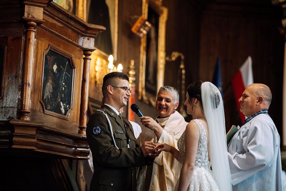 Uroczystość ślubna w kościele: żołnierz w mundurze zakłada obrączkę pannie młodej, błogosławieństwo udzielane przez księdza z mikrofonem, w tle drewniany ołtarz z obrazem i flagi.