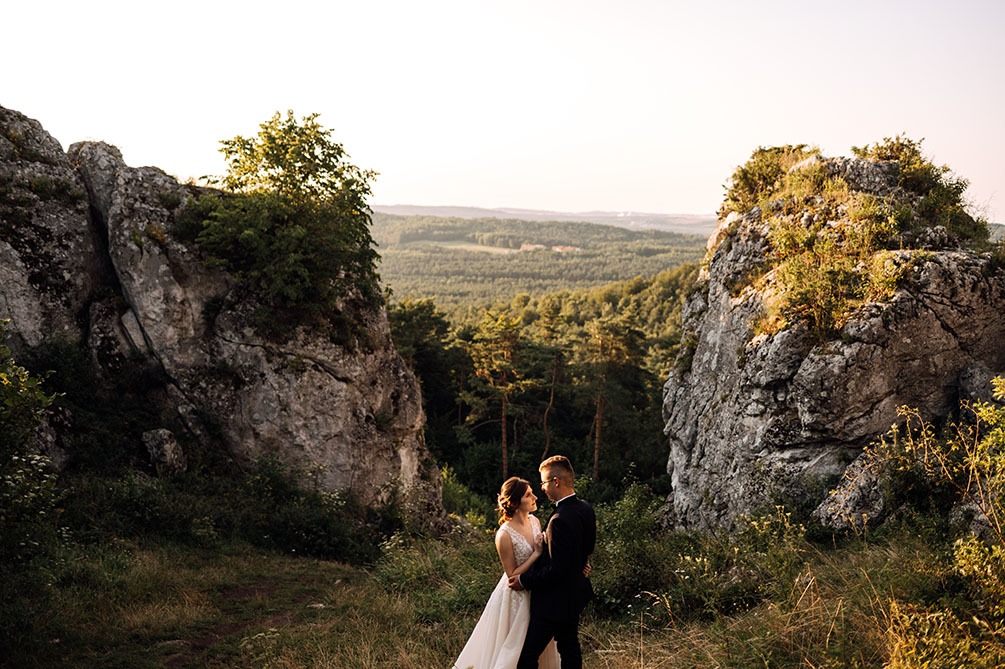 Para młoda w objęciach na tle malowniczego krajobrazu z formacjami skalnymi i lasami w oddali, ciepłe światło zachodzącego słońca.