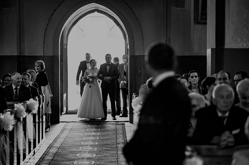 Czarno-białe zdjęcie panny młodej prowadzonej do ołtarza w kościele, z gośćmi po bokach i dywanem w ornamenty na posadzce.