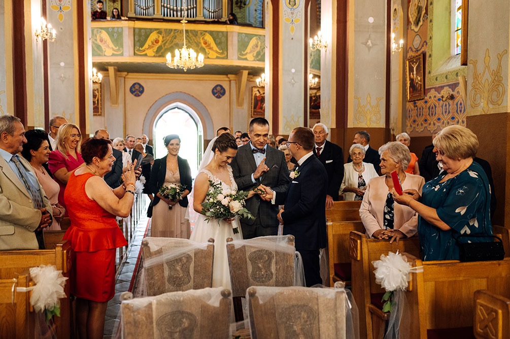 Wymiana obrączek podczas ceremonii ślubnej w kościele, goście obserwują, dekorowane krzesła w pierwszym planie, oświetlenie sufitowe.