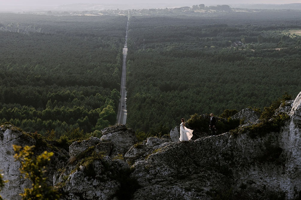 Para młoda na skalistym wzgórzu z widokiem na rozległy las i długą, prostą drogę w oddali.