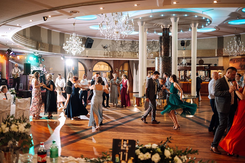 Sala balowa z parkietem, goście weselni tańczą przy muzyce zespołu na scenie, dekoracje kwiatowe na stołach, kryształowe żyrandole.
