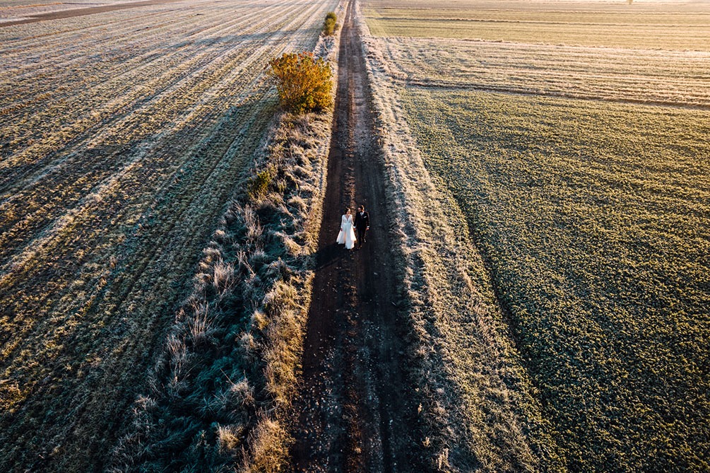 Para młoda spaceruje polną drogą w jesiennej scenerii, widok z drona ukazuje rozległe pola pokryte szronem i rzadkie drzewa w promieniach wschodzącego słońca.