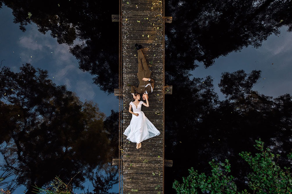 Para młoda leżąca na drewnianym moście nad wodą, widok z góry, w tle odbicie drzew w tafli wody.