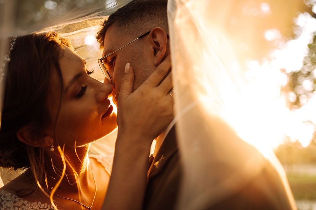 Pocałunek pary młodej pod welonem w kontraście z promieniami zachodzącego słońca, on w okularach i wojskowym mundurze, ona z delikatnym makijażem i perłami.