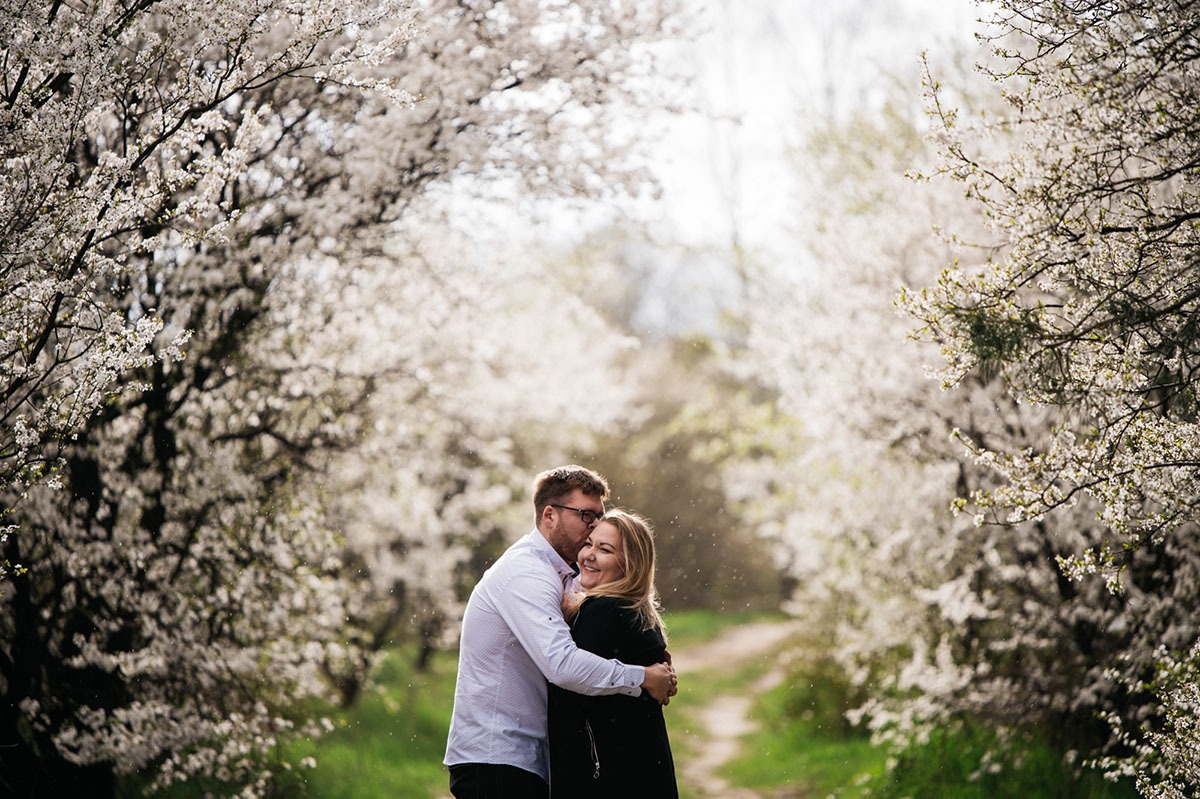 Uśmiechnięta para obejmuje się na tle alei kwitnących drzew w słoneczny dzień, mężczyzna w okularach całuje kobietę w policzek.