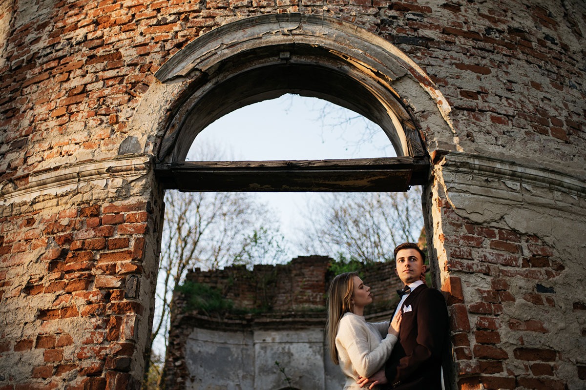 Para młoda pozuje w ruinach ceglanego budynku z łukowym otworem, ona w kremowym swetrze, on w brązowym garniturze z muchą, delikatne światło podkreśla fakturę cegieł.
