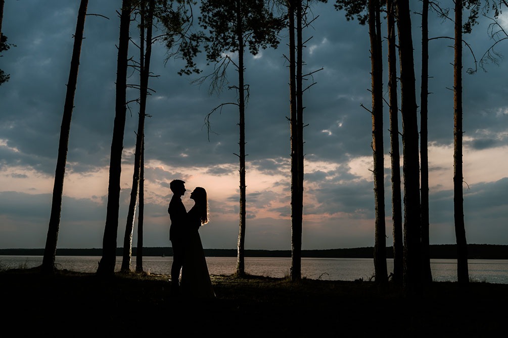 Sylwetki pary młodej na tle jeziora i lasu sosnowego o zmierzchu, delikatne światło podkreśla kontury włosów kobiety.
