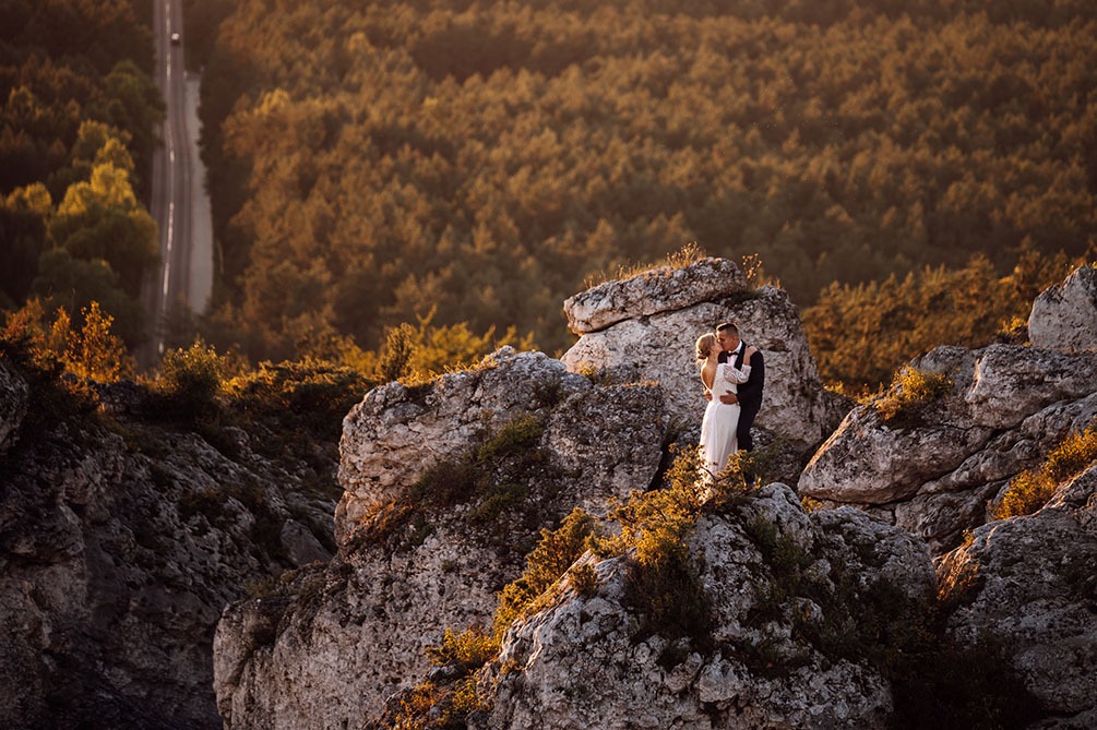Para młoda w objęciach na skalistym wzgórzu, w tle las i droga, ciepłe światło zachodzącego słońca.