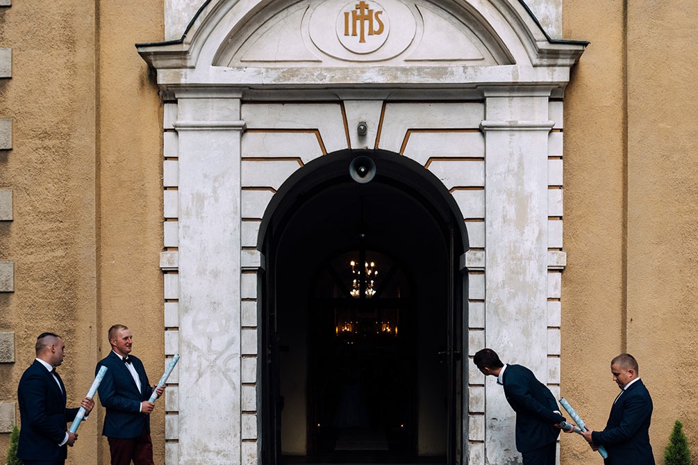 Groom and groomsmen in suits stand at church entrance holding rolled-up papers, preparing for the wedding ceremony.