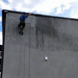 Pracownik zabezpieczony linami myje elewację wysokociśnieniowo, widoczne zacieki na szarej ścianie budynku.
