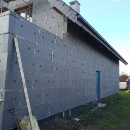 Dom w trakcie prac termoizolacyjnych, ściany pokryte grafitowym styropianem z widocznymi punktami klejenia, rusztowanie w tle, narzędzia i worek na ziemi.