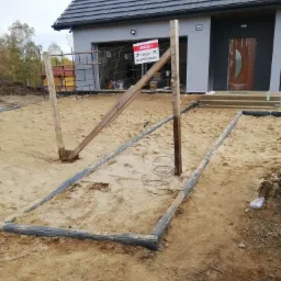 Przygotowanie podłoża pod ułożenie nawierzchni z obrzeżami, widoczny piasek, drewniane słupki i siatka ogrodzeniowa, w tle dom z szarą elewacją i brązowymi drzwiami wejściowymi.