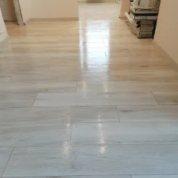 Położona podłoga z jasnych, drewnopodobnych paneli ceramicznych w korytarzu, z widocznymi materiałami budowlanymi w tle.