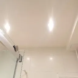 Biała, błyszcząca powłoka sufitu z wbudowanym oświetleniem punktowym nad kabiną prysznicową z elementami armatury i białymi płytkami ściennymi.