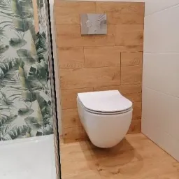 Podwieszana toaleta z chromowanym przyciskiem spłukującym, obudowana płytkami imitującymi drewno, widoczny fragment kabiny prysznicowej z motywem liści na płytkach.