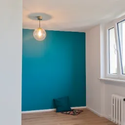 Pomalowane na turkusowo wnętrze pokoju z drewnianą podłogą, białymi ścianami, grzejnikiem żeberkowym, otwartym oknem, poduszką i okrągłą lampą wiszącą.