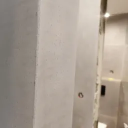 Szpachlowana ściana narożna z widoczną strukturą, w tle częściowo widoczna łazienka w trakcie wykańczania, widoczne elementy instalacji elektrycznej.