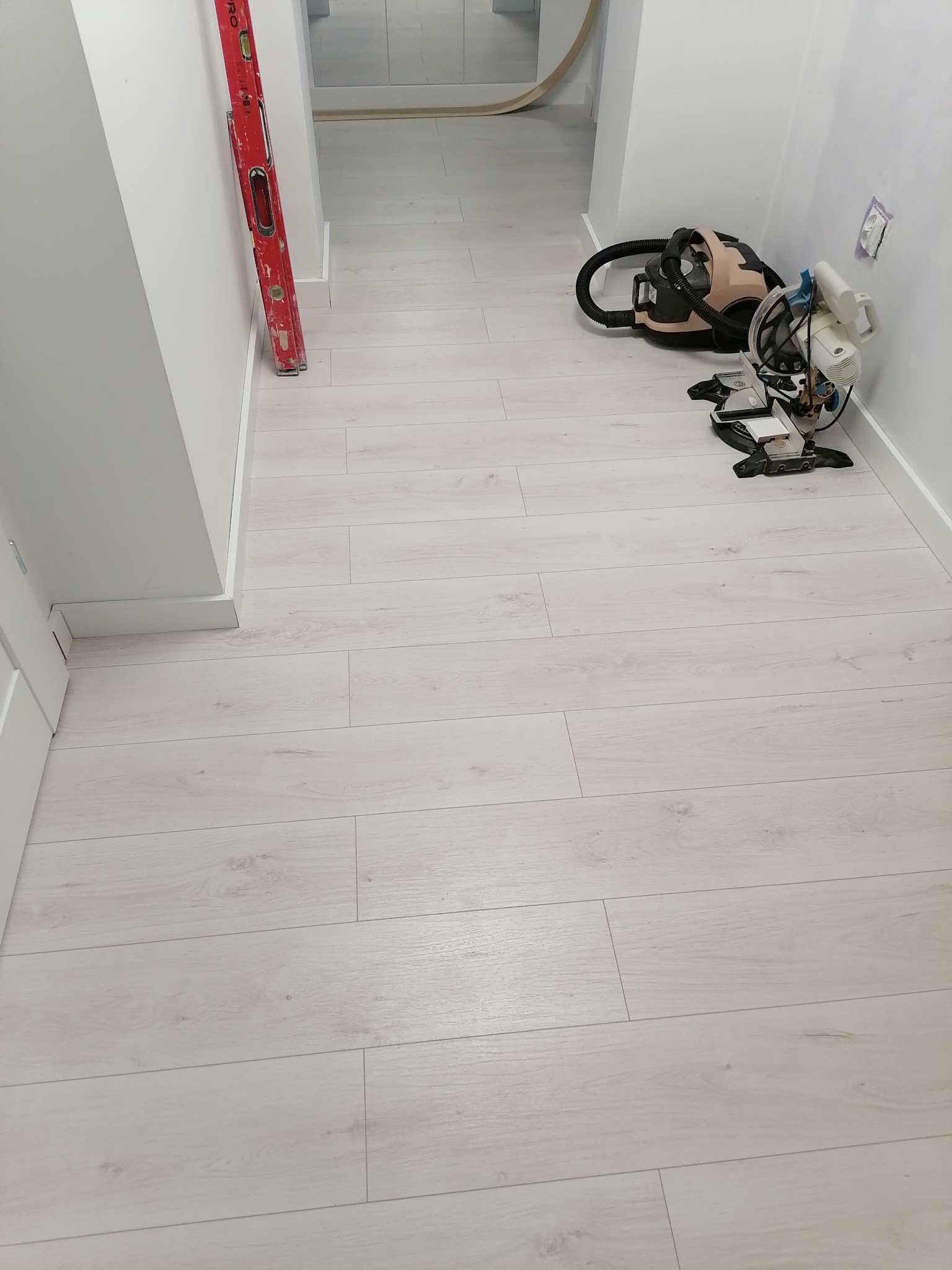 Jasne panele podłogowe ułożone w korytarzu, widoczna poziomica oparta o ścianę, odkurzacz i piła do paneli przy ścianie, listwy przypodłogowe zamontowane.