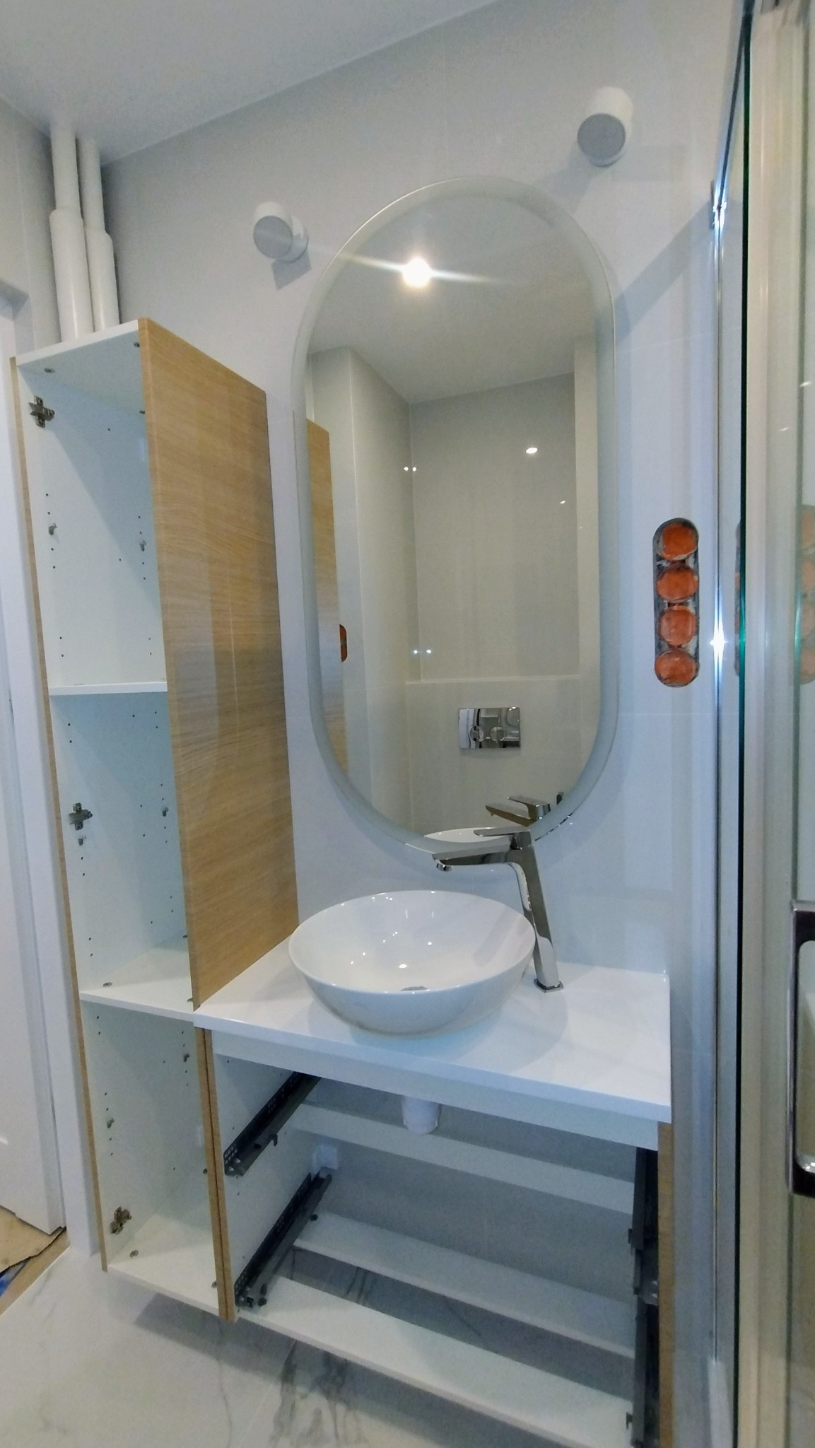 Nowoczesna łazienka w trakcie montażu: biała umywalka nablatowa z chromowaną baterią, owalne lustro z podświetleniem, szafka z drewnianym frontem i otwartymi półkami w trakcie instalacji.