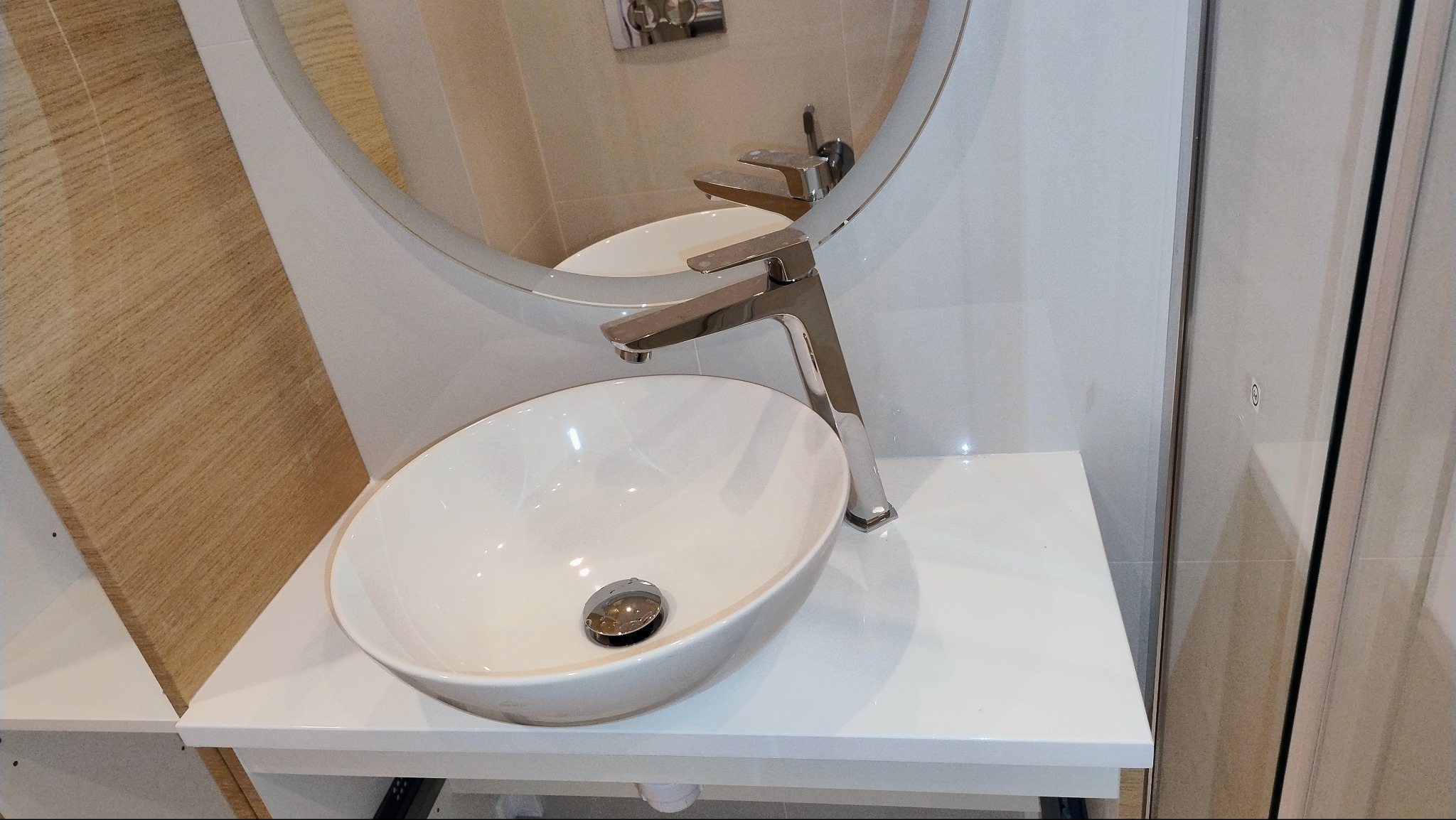 Biała, owalna umywalka nablatowa z chromowaną baterią, zamontowana na białym blacie w nowoczesnej łazience z okrągłym lustrem.