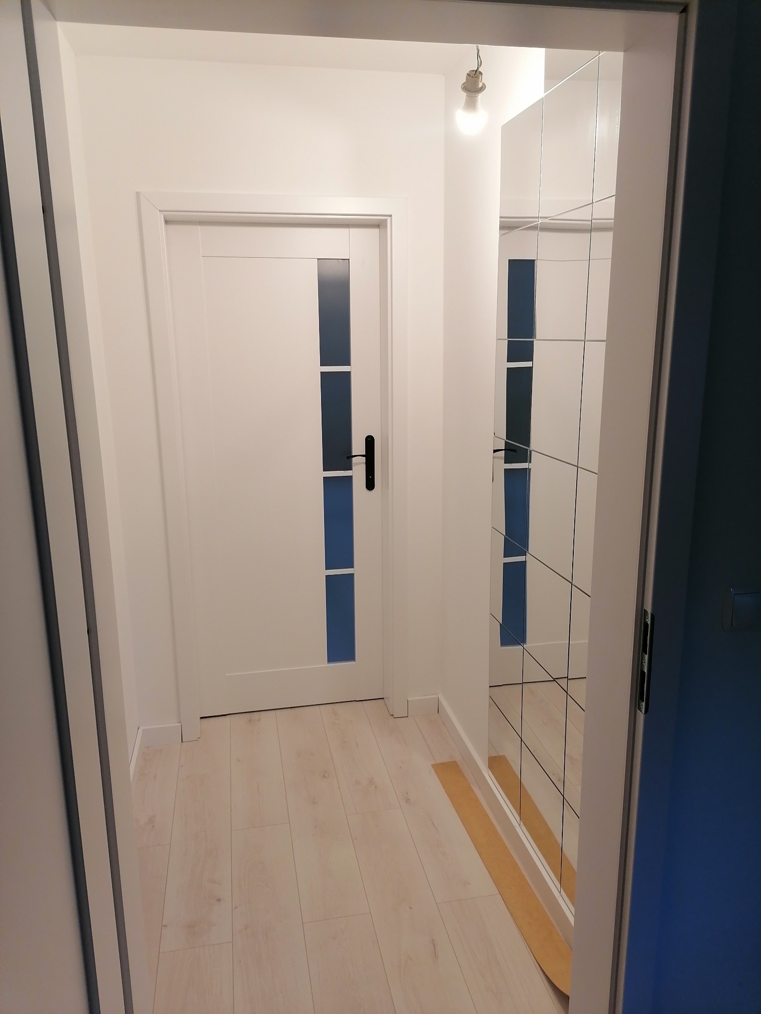 Korytarz po remoncie: białe ściany, panele podłogowe w odcieniach beżu, białe drzwi z przeszkleniami w kolorze niebieskim i duże lustro w panelach na ścianie.