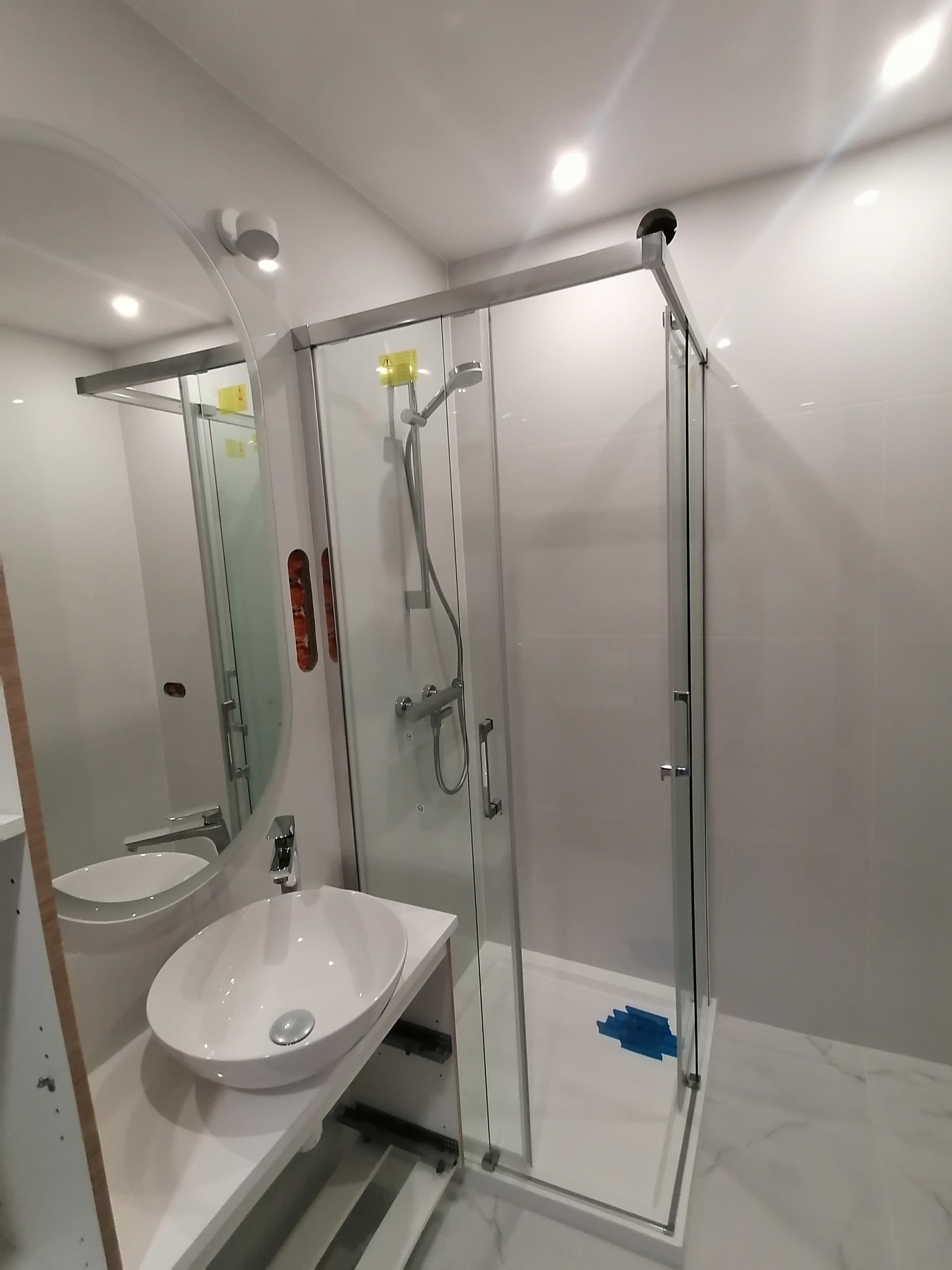 Nowoczesna łazienka z kabiną prysznicową z przesuwnymi drzwiami, owalną umywalką nablatową i dużym okrągłym lustrem, widoczne zabezpieczenia folią na brodziku.