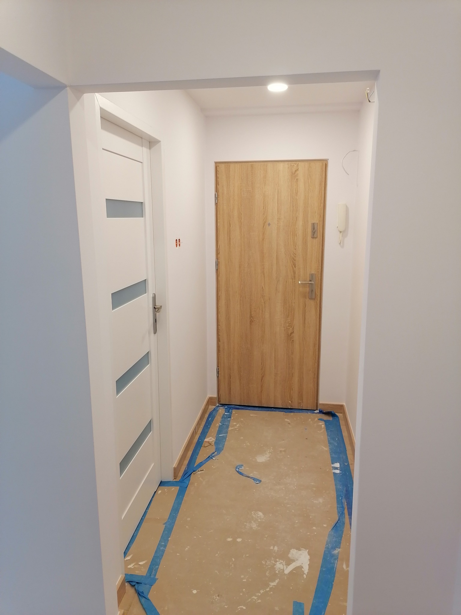 Korytarz w trakcie prac wykończeniowych, zabezpieczona podłoga folią i taśmą malarską, widoczne dwie pary drzwi: białe wewnętrzne z przeszkleniami i drewnopodobne zewnętrzne.